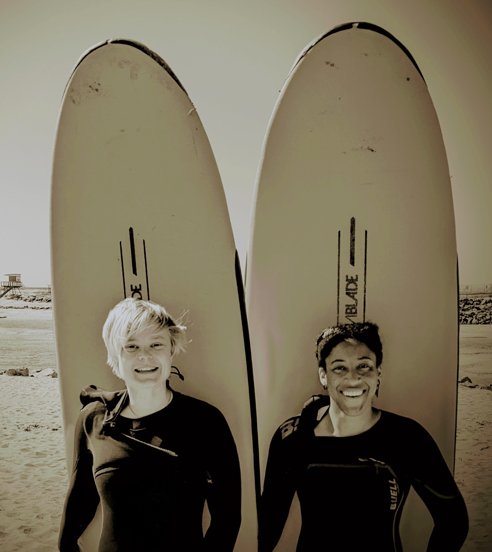 Kendra and companion in wetsuits in front of surfboards